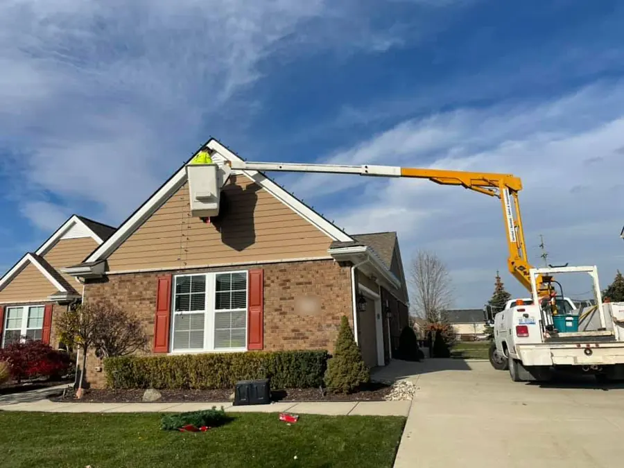 A crew member in a bucket lift at the peak of a brick and sided home, with a white utility truck in the driveway.