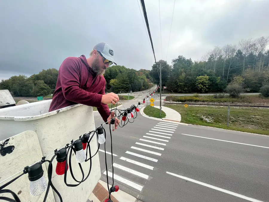 A crew member in a bucket lift attaching a strand of red and clear bistro bulbs to an overhead cable above a road crosswalk.