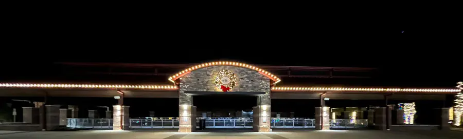 A covered pavilion at night with warm white bulbs outlining the roofline and a stone center gable holding a lit wreath.
